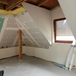 WNB Decor - Poddasze w trakcie prac wykończeniowych: zamontowane płyty na skośnych ścianach, widoczne drewniane belki stropowe, okno z roletą wewnętrzną, podłoga przygotowana do dalszych prac.