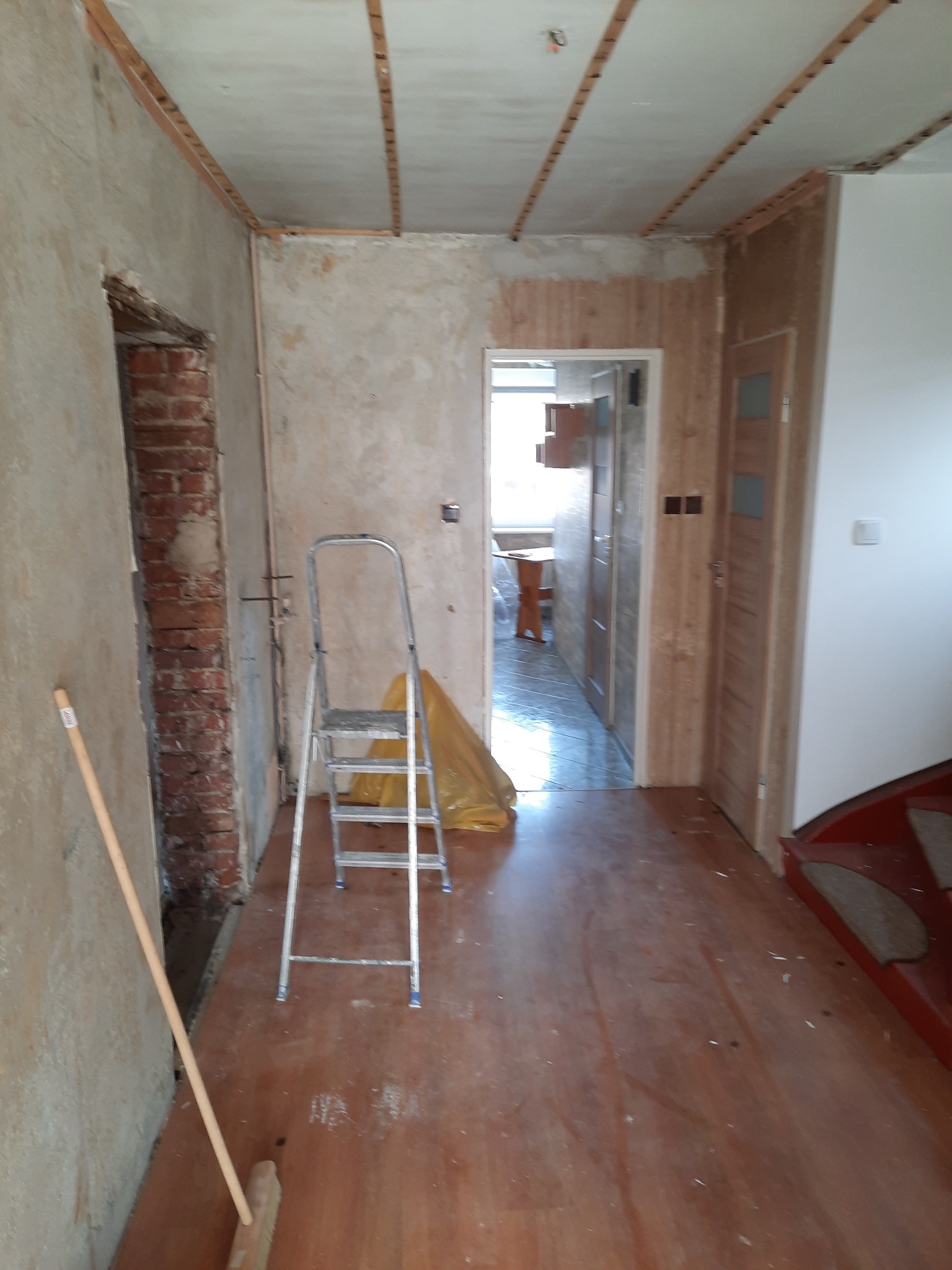 Wnętrze mieszkania w trakcie remontu: ściany w stanie surowym, odsłonięta cegła, drewniane listwy na suficie, aluminiowa drabina i częściowo wykończona podłoga z widocznymi schodami.