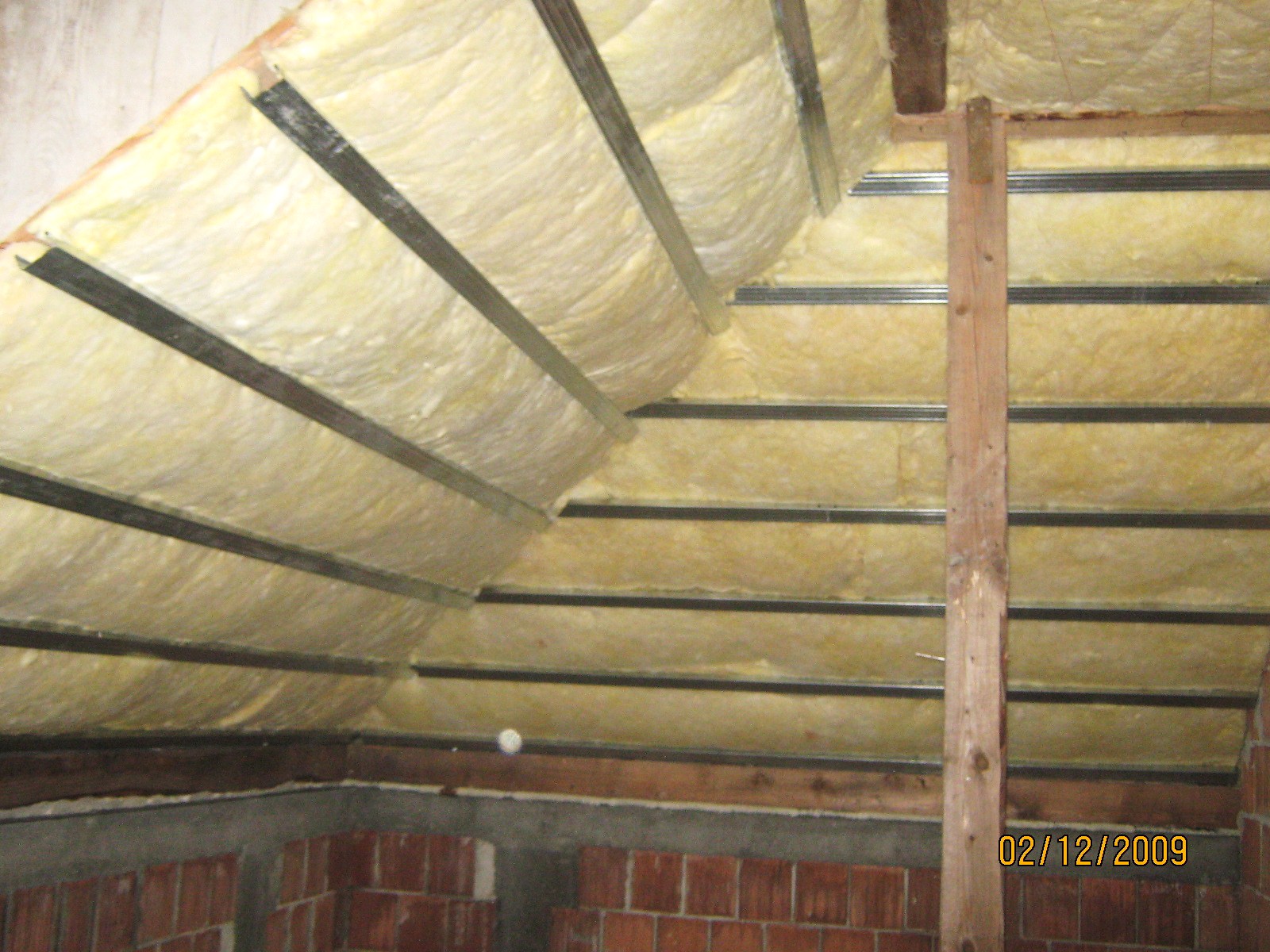 Izolacja poddasza wełną mineralną, widoczne metalowe profile konstrukcyjne, ceglany fragment ściany w dolnej części kadru, data wykonania zdjęcia w prawym dolnym rogu.