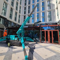 AIRON PRO-TECH Sławomir Kos - Montaż aluminiowych okien i drzwi w nowoczesnym budynku. Użycie miniżurawia. Pracownicy w kaskach i kamizelkach. Widoczne elementy konstrukcyjne. Jasne, dzienne światło.