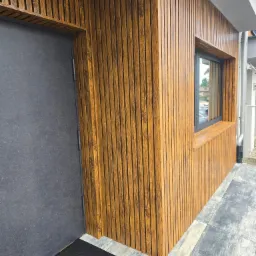 Elewacja budynku z lamelami w odcieniach brązu i szara, minimalistyczna stolarka okienna oraz drzwi wejściowe. Nowoczesny design.