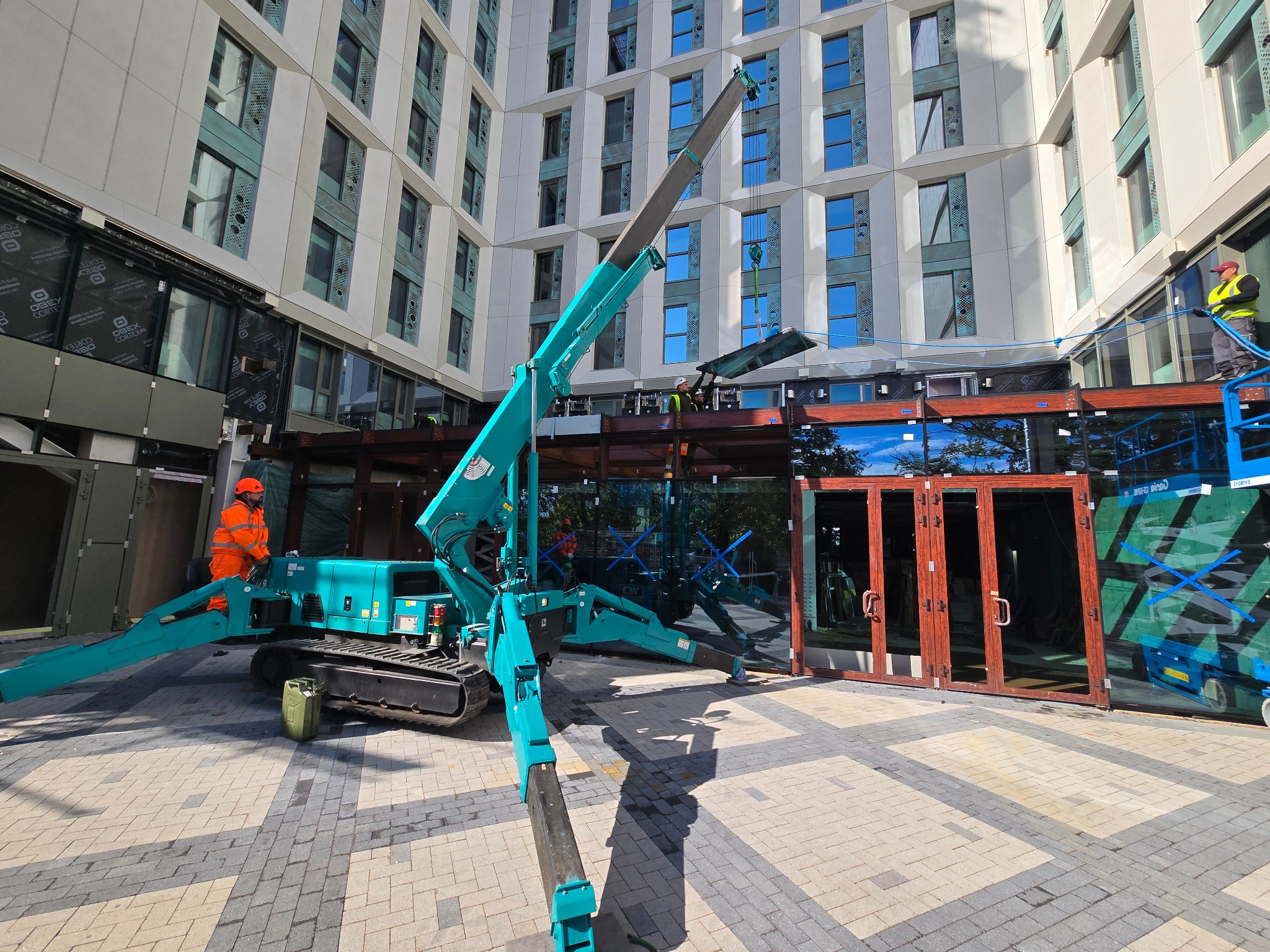 Montaż aluminiowych okien i drzwi w nowoczesnym budynku. Użycie miniżurawia. Pracownicy w kaskach i kamizelkach. Widoczne elementy konstrukcyjne. Jasne, dzienne światło.