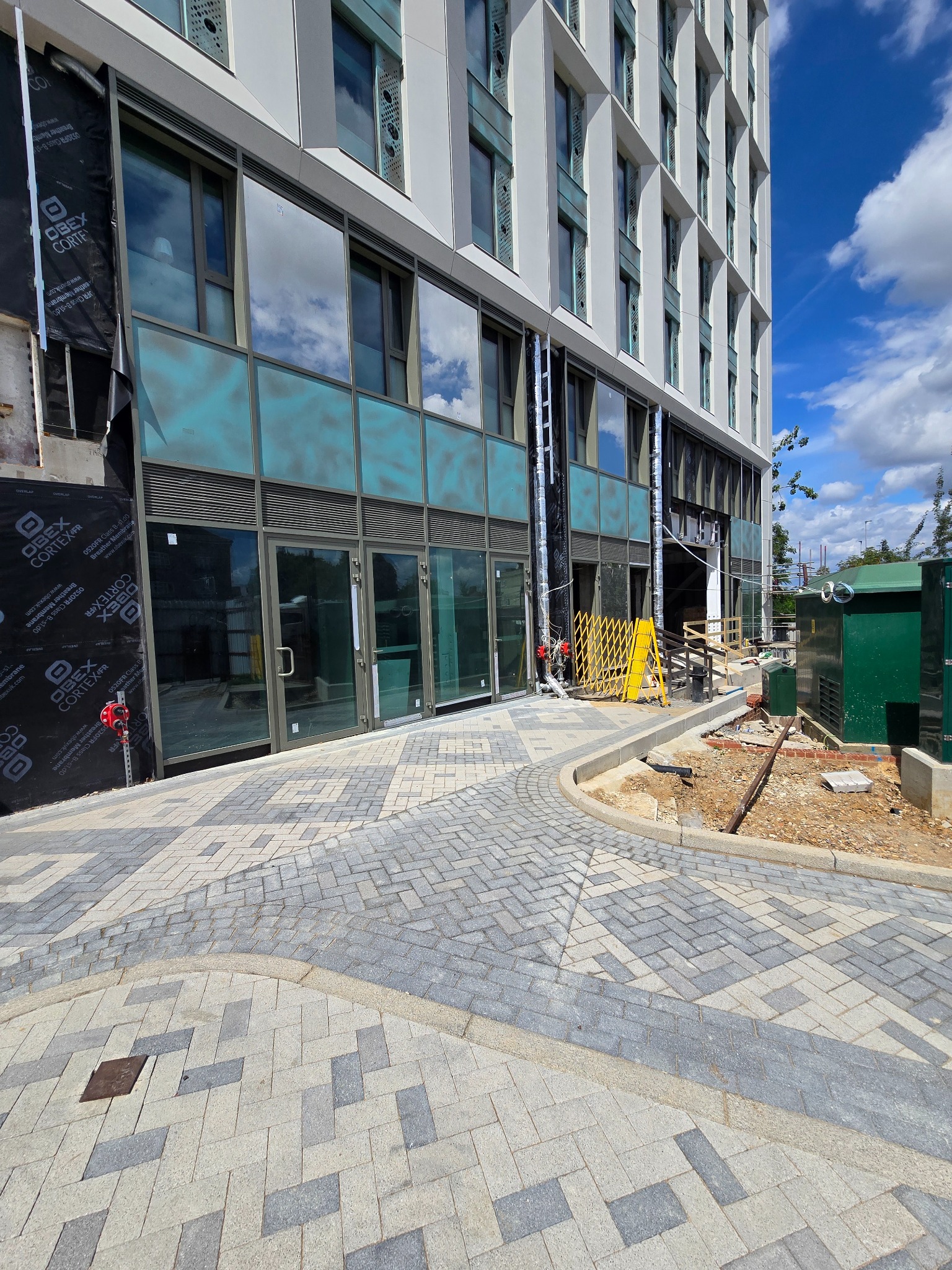 Budynek z aluminiowymi oknami podczas montażu; widoczne rusztowania i zabezpieczenia. Na pierwszym planie ułożona kostka brukowa.