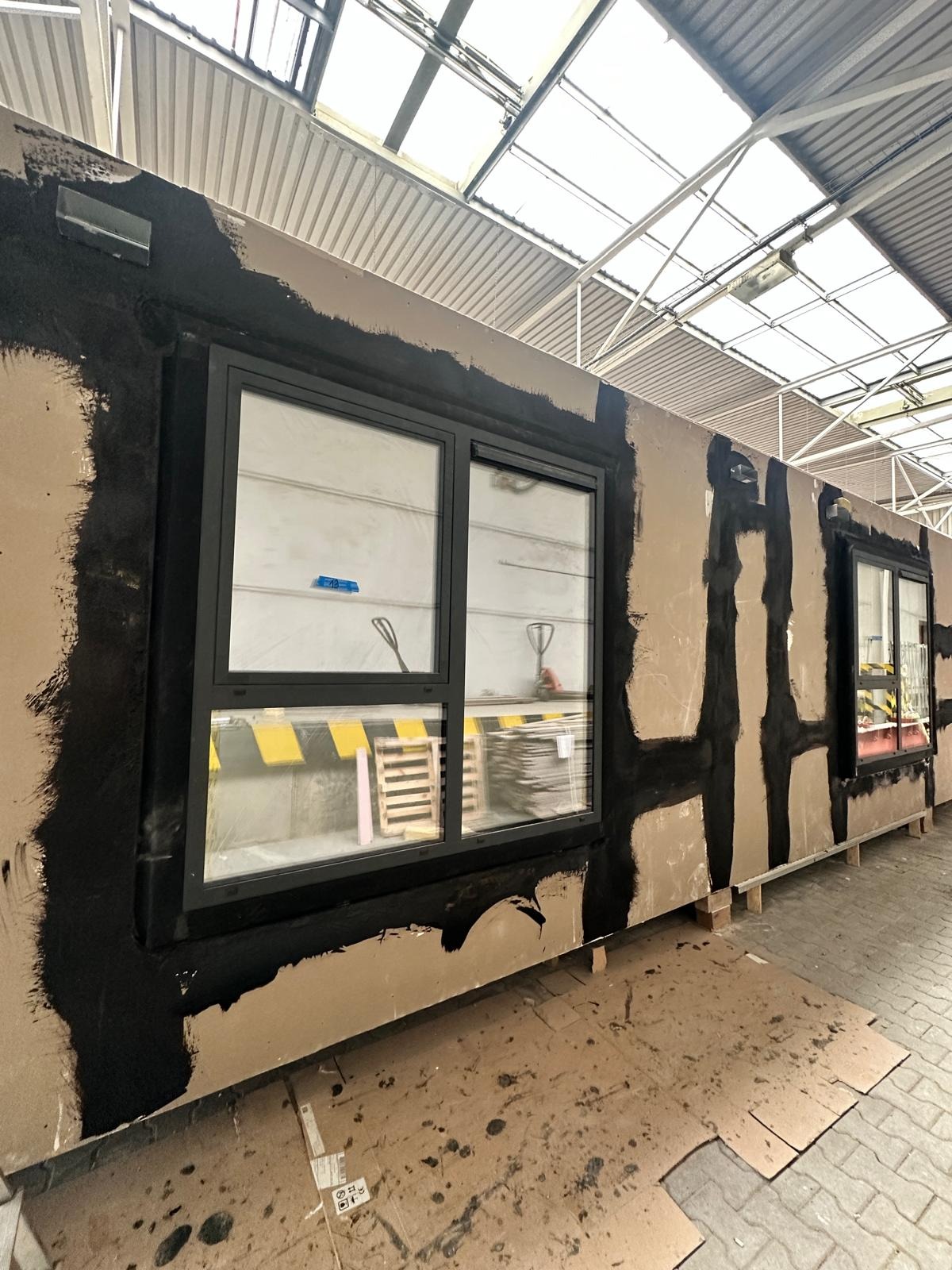 Montaż okna aluminiowego w surowym otworze. Widoczne czarne uszczelnienie i przygotowanie do dalszych prac wykończeniowych. W tle palety i elementy konstrukcyjne.