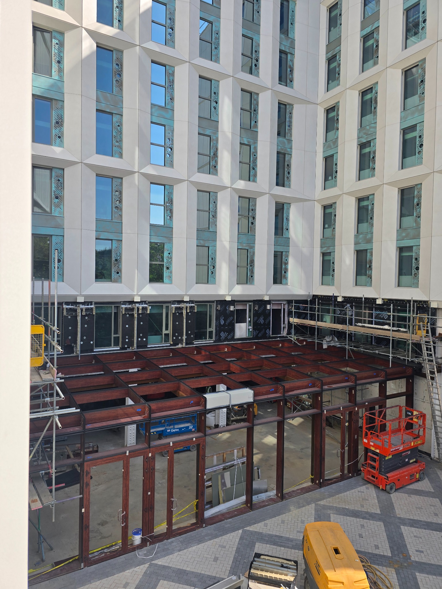 Montaż stolarki okiennej i drzwiowej w budynku o nowoczesnej architekturze. Widoczne rusztowania, podnośniki i elementy konstrukcyjne.