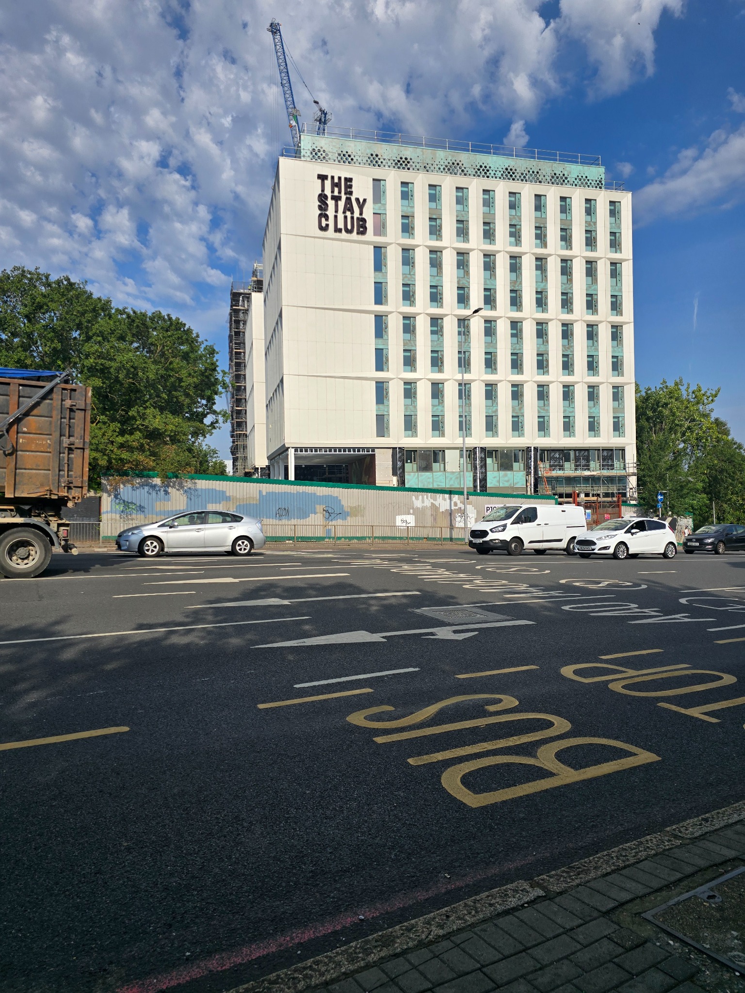 Budynek 'The Stay Club' z widocznymi oknami aluminiowymi, częściowo w rusztowaniach. Na pierwszym planie ulica z oznakowaniem 'BUS STOP'. Błękitne niebo z chmurami w tle.