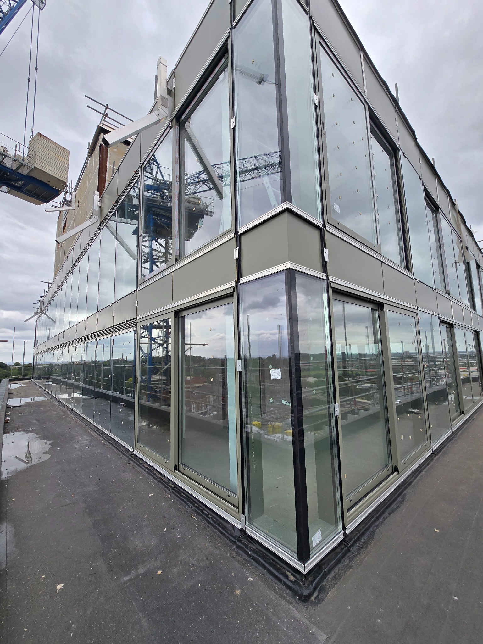 Szklana fasada budynku w trakcie montażu, widoczne ramy okienne i elementy konstrukcyjne. Na pierwszym planie fragment dachu. W tle dźwig budowlany.
