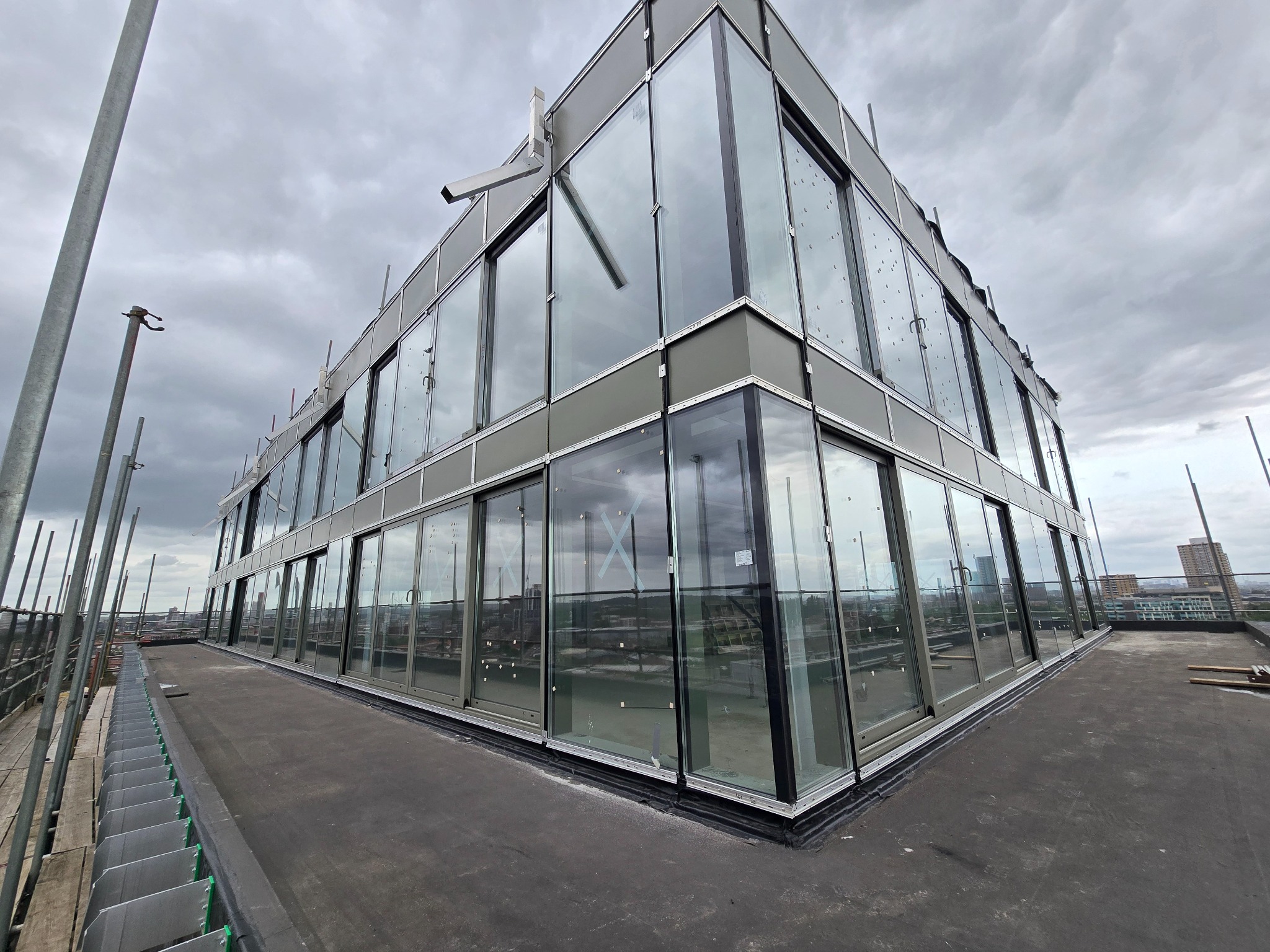 Nowoczesny budynek z szklaną fasadą na dachu, widok z zewnątrz, szare niebo w tle. Widoczne elementy rusztowania, co sugeruje trwające prace wykończeniowe.