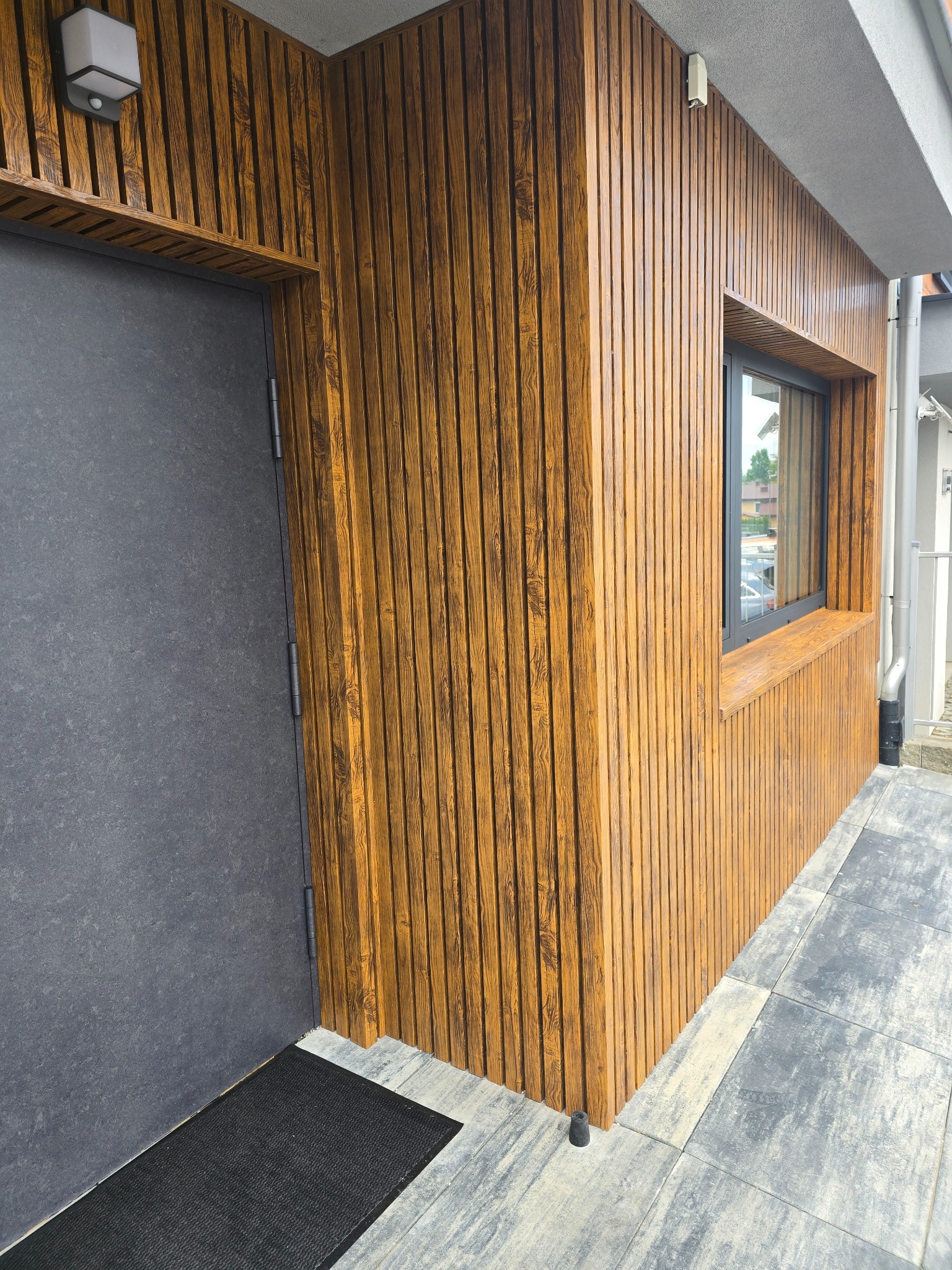 Elewacja budynku z lamelami w odcieniach brązu i szara, minimalistyczna stolarka okienna oraz drzwi wejściowe. Nowoczesny design.