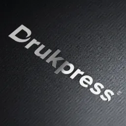 Srebrny napis 'Drukpress' na ciemnoszarym, teksturowanym tle z delikatnym wzorem prążków.