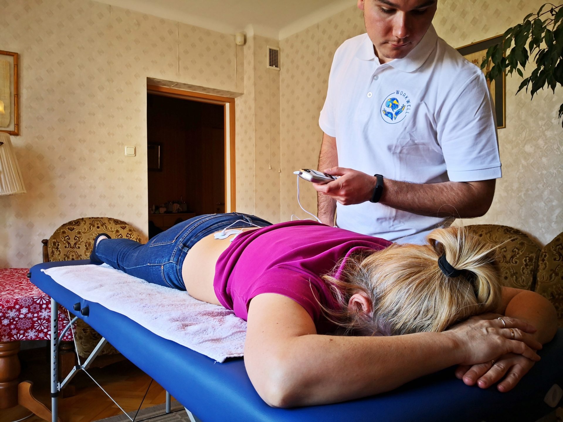 Pacjentka leży na niebieskim stole do masażu, podłączona elektrodami do urządzenia trzymanego przez mężczyznę w białej koszulce z logo 'WORK WELL'.