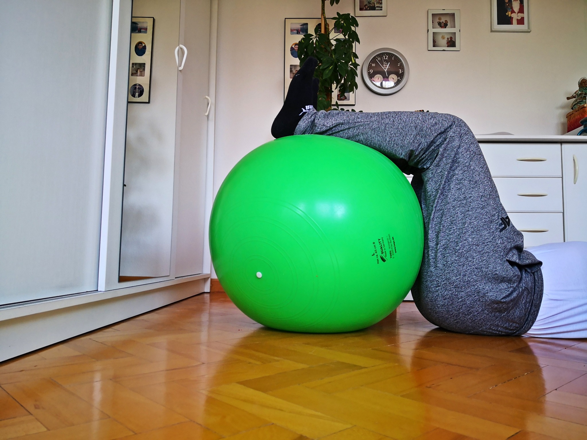 Osoba w szarych spodniach i czarnej skarpecie wykonuje ćwiczenie z zieloną piłką rehabilitacyjną w domowym otoczeniu, na parkiecie, z widocznymi meblami i dekoracjami na ścianie.