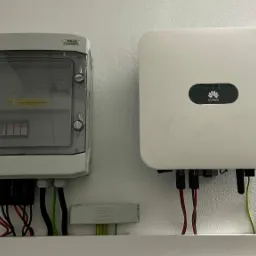 Biały inwerter Huawei i skrzynka zabezpieczeń KENO z przewodami na białej ścianie w Sosnowcu, element instalacji.