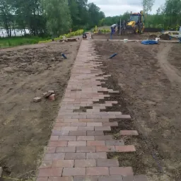 Układanie czerwonej kostki brukowej na ścieżce w ogrodzie, widoczny fragment ułożonej nawierzchni, przygotowany teren i pracownicy z ciężkim sprzętem w tle.