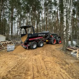 Czerwono-czarny traktor z przyczepą marki Pronar, wypełnioną piaskiem, stoi na piaszczystym terenie z widocznymi śladami opon, w tle las mieszany z przewagą drzew iglastych i brzozy.