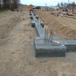 Wylane betonowe fundamenty z wystającymi prętami zbrojeniowymi na placu budowy, widoczni pracownicy w odblaskowych kamizelkach oraz samochody w tle.