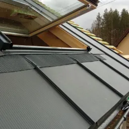 Fragment dachu z otwartym oknem dachowym i widocznymi elementami konstrukcyjnymi, w tle drzewa.