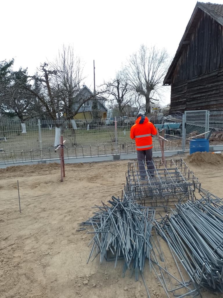 Pracownik w pomarańczowej kurtce stoi obok zbrojenia fundamentów na placu budowy, widoczne elementy szalunku i przygotowane pręty.