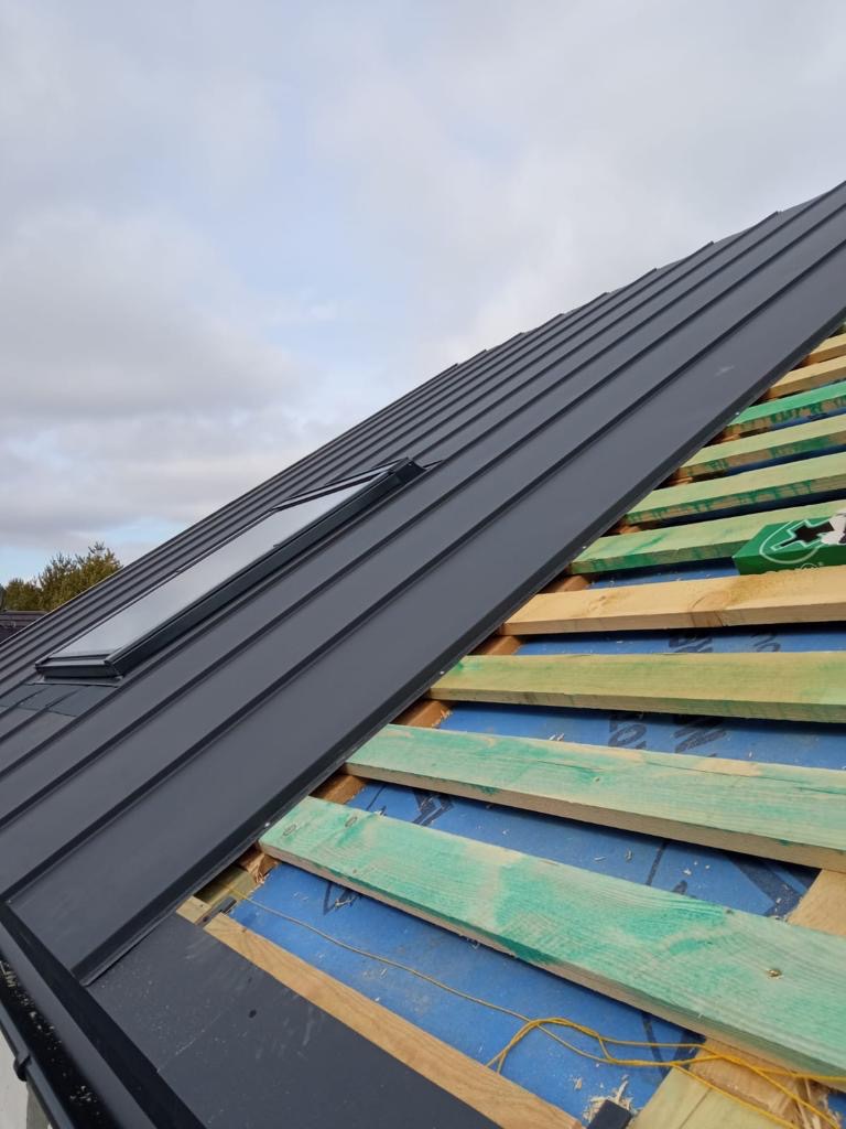 Montaż dachu z blachy na drewnianej konstrukcji z oknem dachowym, widoczne elementy izolacji i niebieska membrana dachowa.