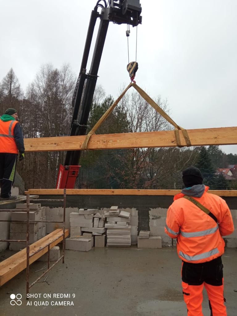 Montaż drewnianego elementu konstrukcyjnego z użyciem dźwigu na budowie domu z bloczków betonowych; pracownicy w odblaskowych ubraniach nadzorują proces.
