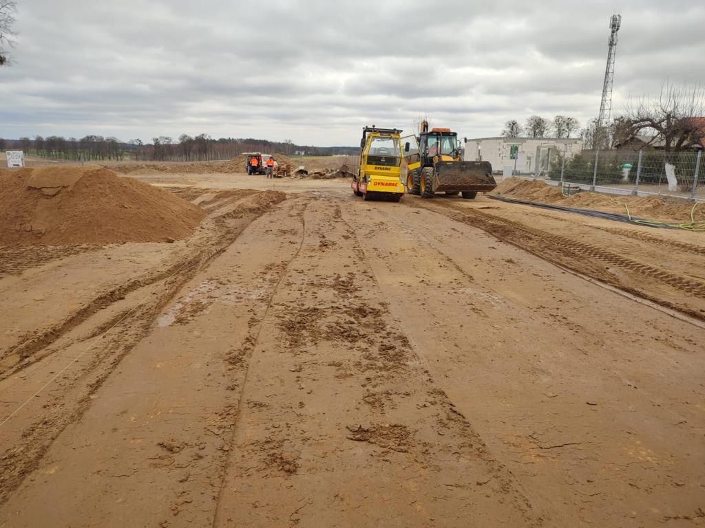 Przygotowanie terenu pod budowę: ubijarka wibracyjna Dynapac i koparko-ładowarka na błotnistym placu budowy, w tle sterta ziemi i budynek w trakcie wznoszenia.