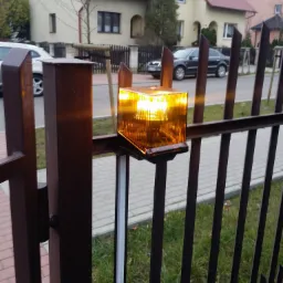 Pomarańczowa lampa ostrzegawcza zamontowana na metalowym słupku bramy wjazdowej; w tle widoczne domy jednorodzinne i zaparkowane samochody.