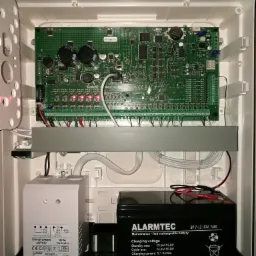 Wnętrze obudowy centrali alarmowej z widoczną płytą główną, transformatorem i akumulatorem podtrzymującym; komponenty elektroniczne i okablowanie.
