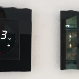 Dwa czarne, dotykowe panele sterowania na białej ścianie. Jeden wyświetla temperaturę 23 stopnie Celsjusza, drugi ma okrągły przycisk dotykowy. Widoczne odbicie osoby w panelu z wyświetlaczem.