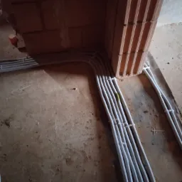 Rury instalacyjne w korytach ściennych z cegły dziurawki, zabezpieczone opaskami zaciskowymi, widok z góry na surową podłogę.