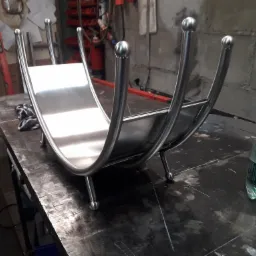 Metalowy stojak na drewno kominkowe z polerowanej stali nierdzewnej, z zaokrąglonymi bokami zakończonymi kulkami, leżący na warsztatowym stole obok butelki wody.