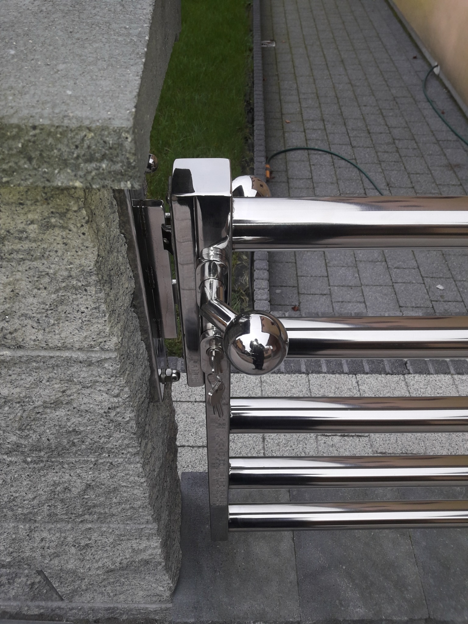 Fragment ogrodzenia ze stali nierdzewnej z kulistą gałką i kluczykiem przy bramie w Kiełpinie, widoczne mocowanie do kamiennego słupka.