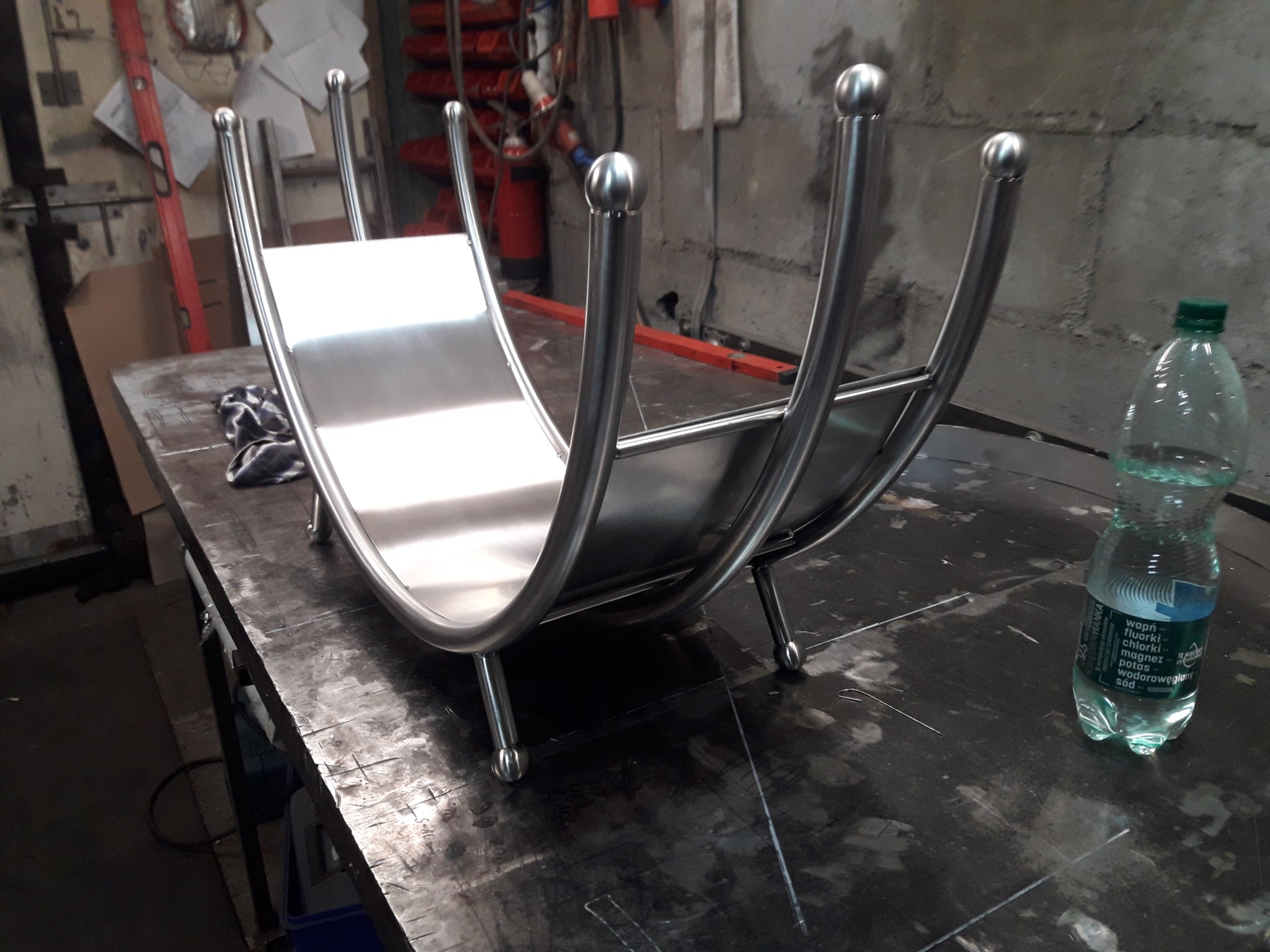 Metalowy stojak na drewno kominkowe z polerowanej stali nierdzewnej, z zaokrąglonymi bokami zakończonymi kulkami, leżący na warsztatowym stole obok butelki wody.