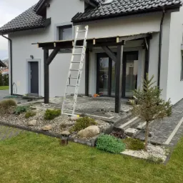 Dom jednorodzinny z szarą elewacją, czarną stolarką okienną i zadaszeniem tarasu w trakcie budowy, drabina oparta o zadaszenie, ułożona częściowo kostka brukowa na tarasie, zagospodarowany ogród...