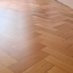 Parkiet ułożony w jodełkę, częściowo wycyklinowany, widoczne różnice w odcieniu drewna, ściany w kolorze ecru, grzejnik przy podłodze.