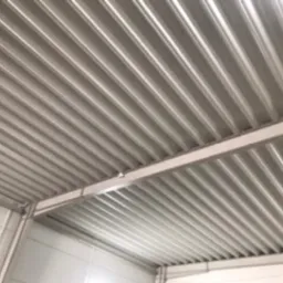 Metalowa konstrukcja dachu hali magazynowej z widocznymi panelami blachy trapezowej, oświetlona sztucznym światłem.