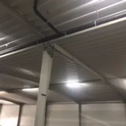 Wnętrze hali magazynowej w trakcie budowy, widoczne stalowe elementy konstrukcyjne, blaszane poszycie dachu i instalacja prowadzona pod stropem.