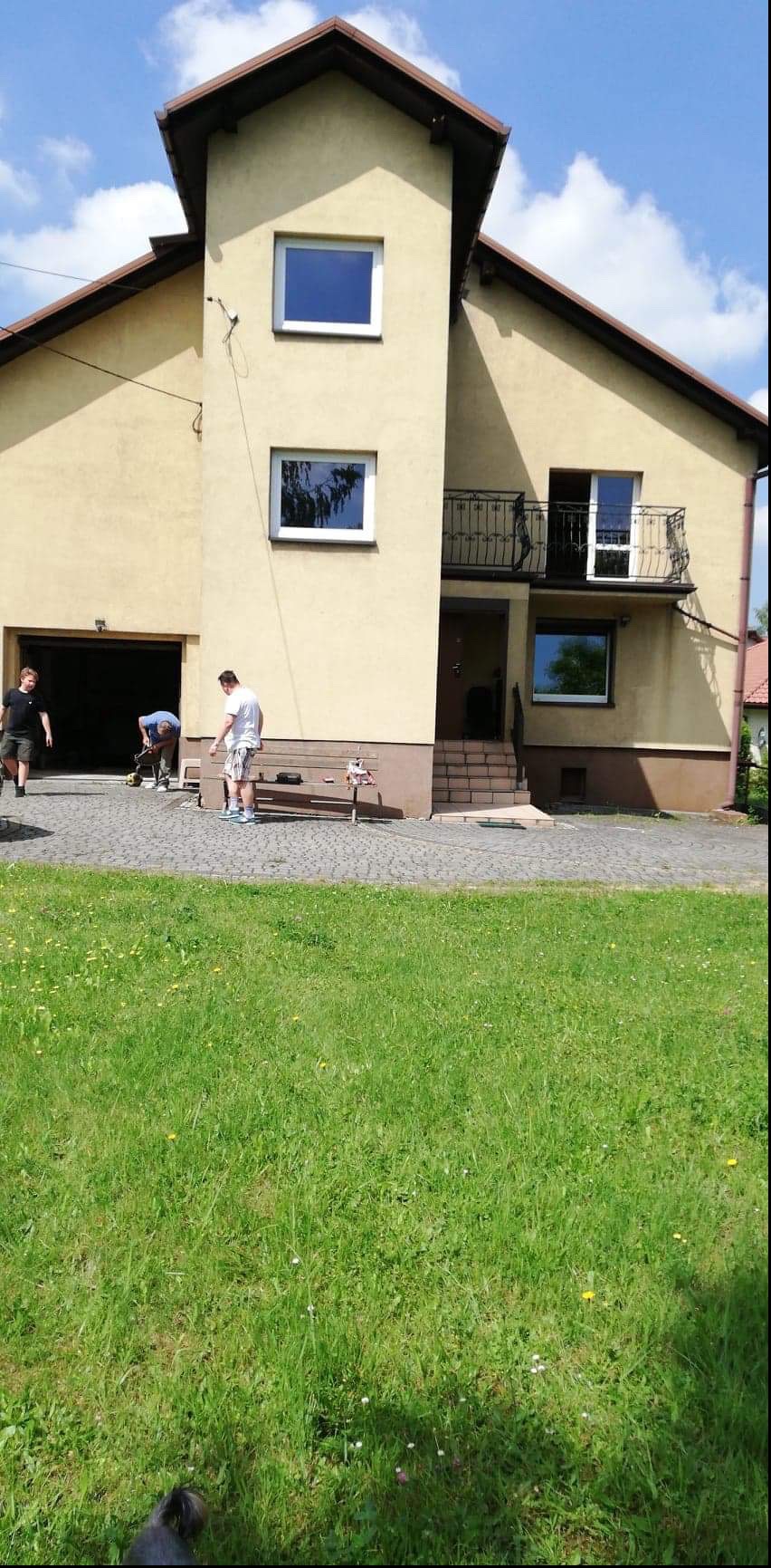 Prace brukarskie przed domem z balkonem, widoczni trzej mężczyźni podczas układania kostki brukowej na podjeździe, narzędzia na ławce.