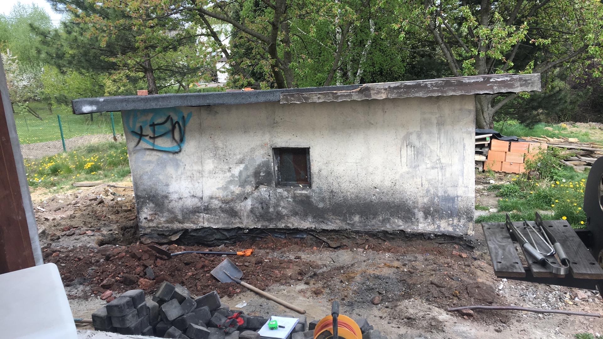 Stary, betonowy budynek gospodarczy z graffiti i małym okienkiem z kratą, przygotowywany do remontu, widoczne narzędzia i materiały budowlane.
