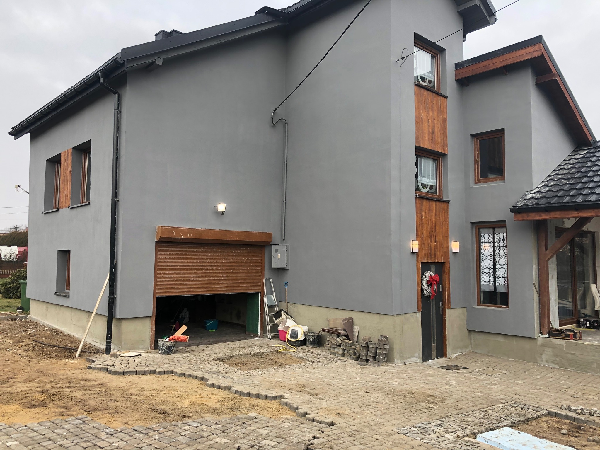 Szary dom jednorodzinny z drewnianymi elementami wykończeniowymi, częściowo ułożona kostka brukowa na podjeździe, wieniec świąteczny na drzwiach wejściowych.