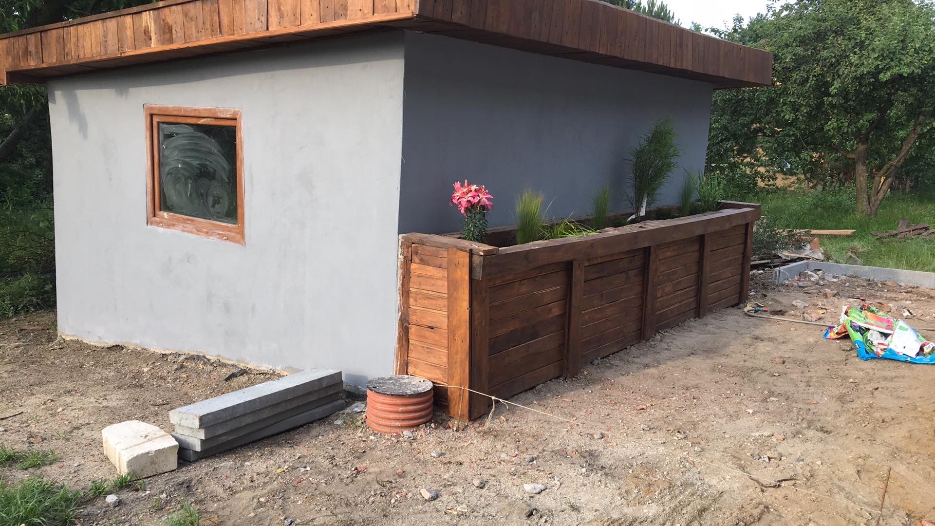Nowy budynek z szarą elewacją, drewnianym oknem i dachem, z drewnianą rabatą kwiatową, na ziemi leżą betonowe bloczki.