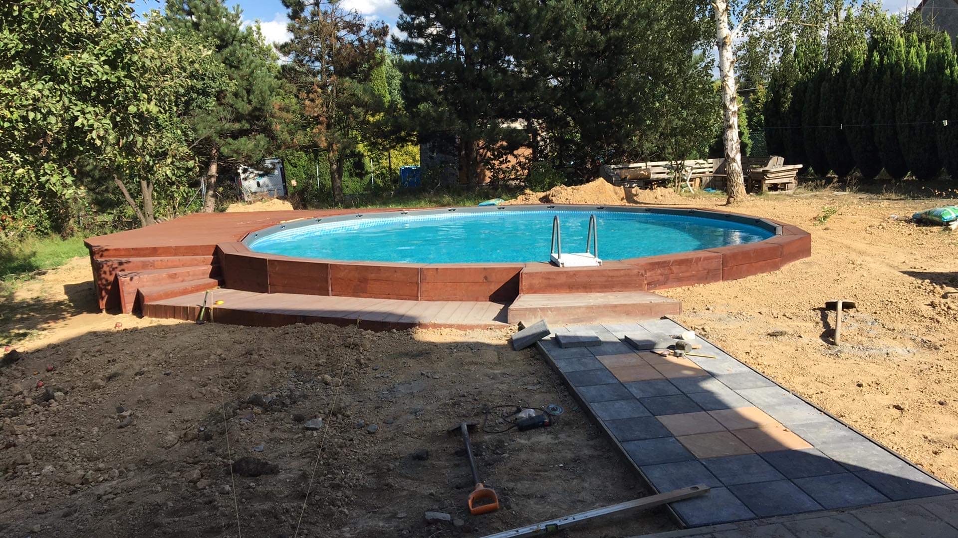 Owalny basen ogrodowy z drewnianą obudową i schodkami w trakcie budowy, widoczne narzędzia, częściowo ułożona kostka brukowa, ziemia wokół.