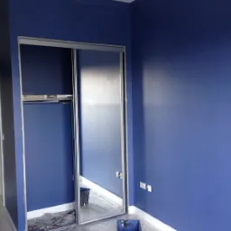 Pokój po malowaniu na ciemny odcień niebieskiego, z zabezpieczoną folią podłogą i widocznymi puszkami z farbą oraz narzędziami malarskimi przy ścianie, z odsuwanymi drzwiami szafy wnękowej.