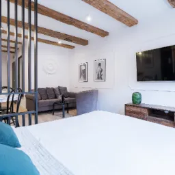 Nowoczesne wnętrze apartamentu z widokiem na salon i jadalnię przez metalową ażurową ściankę działową, drewniane belki sufitowe, telewizor na ścianie i dekoracyjne sztukaterie.