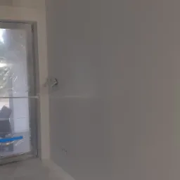 Pracownik w masce ochronnej aplikuje farbę natryskowo na ścianę w pomieszczeniu, okno zabezpieczone folią, widoczne puszki z farbą.