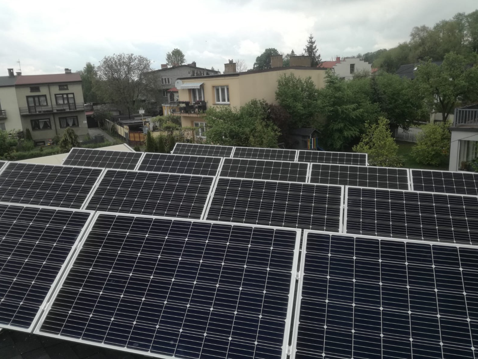 Rząd paneli słonecznych na dachu budynku mieszkalnego, widok z góry, w tle zabudowa miejska i zieleń.