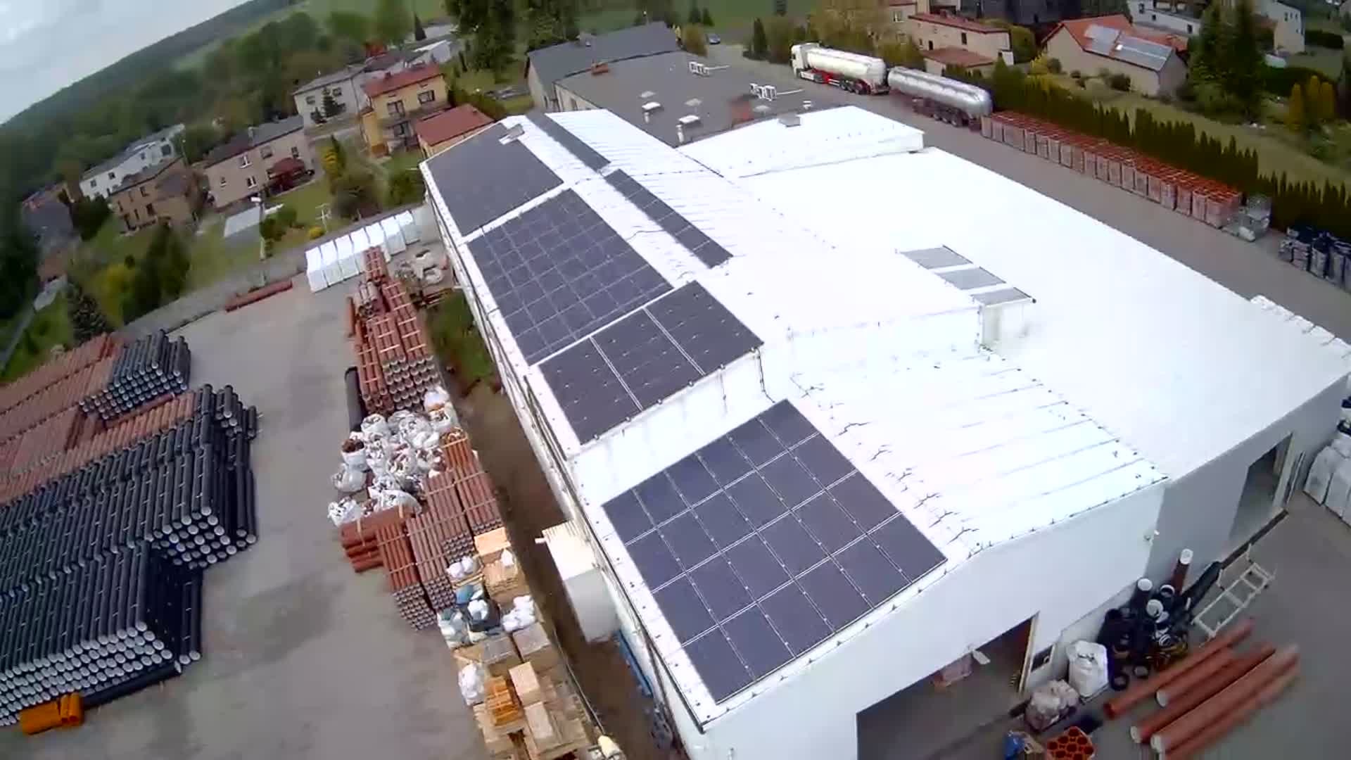 Widok z góry na biały budynek przemysłowy z zamontowanymi panelami słonecznymi na dachu, obok składowane rury i materiały budowlane, w tle zabudowania mieszkalne i tereny zielone.
