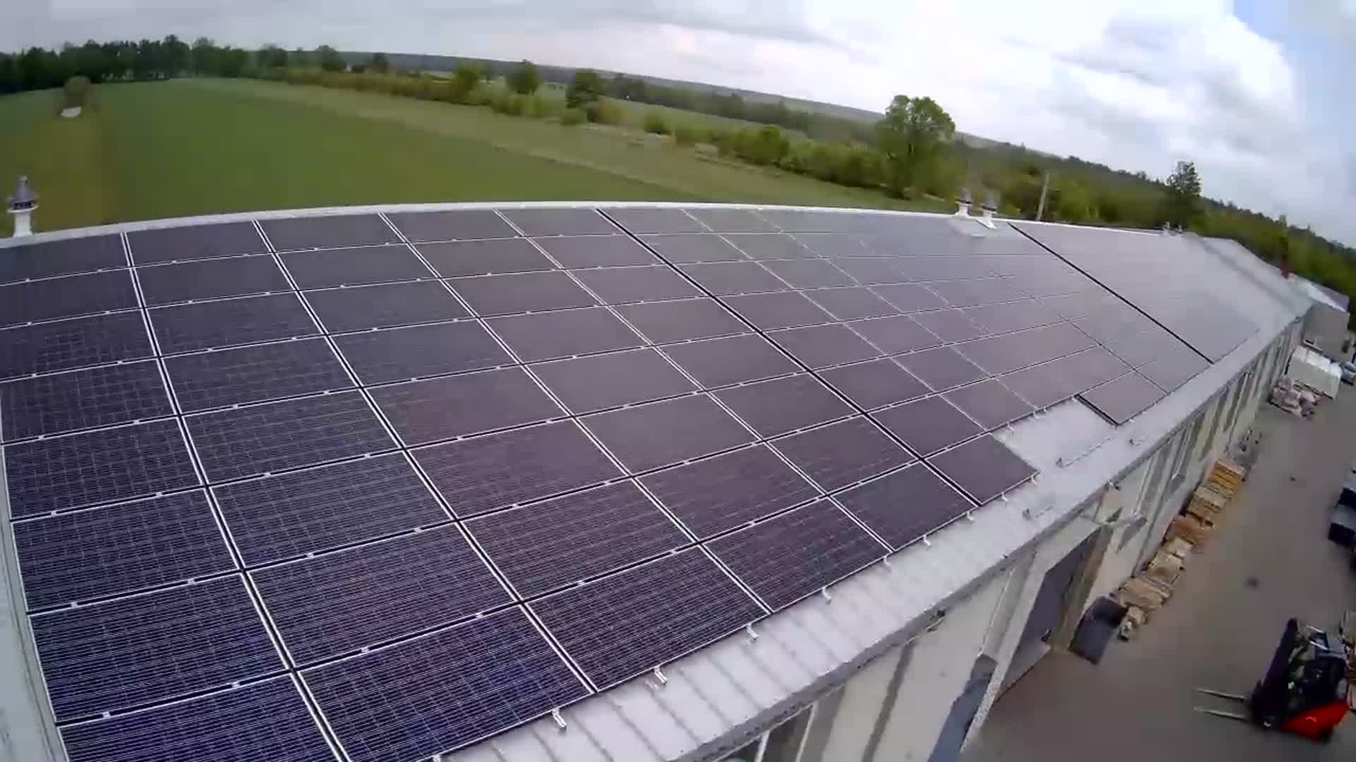Rozległa instalacja paneli słonecznych na dachu budynku przemysłowego, widok z góry, z widocznym w tle krajobrazem polnym i leśnym.