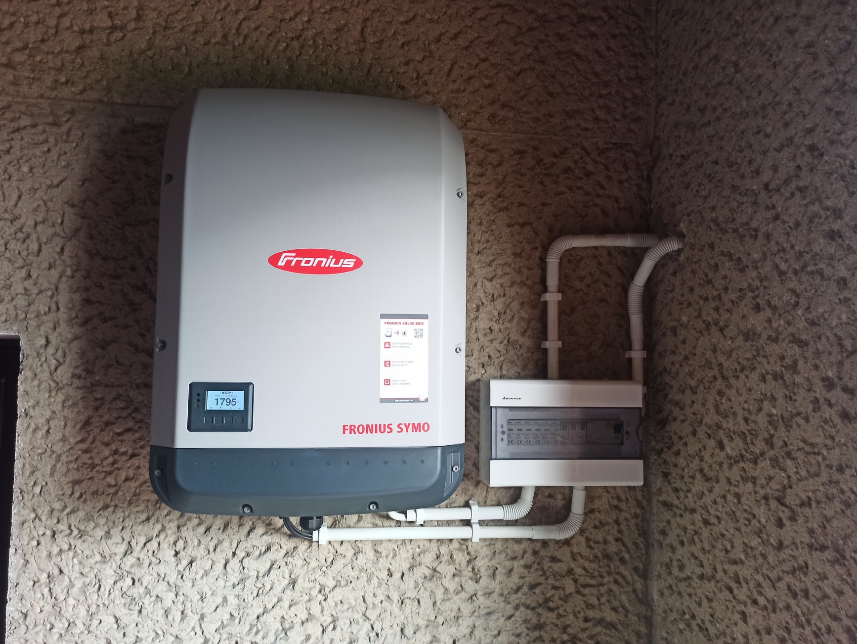Szary inwerter Fronius Symo zamontowany na teksturowanej ścianie, obok skrzynka elektryczna z białymi rurkami ochronnymi.
