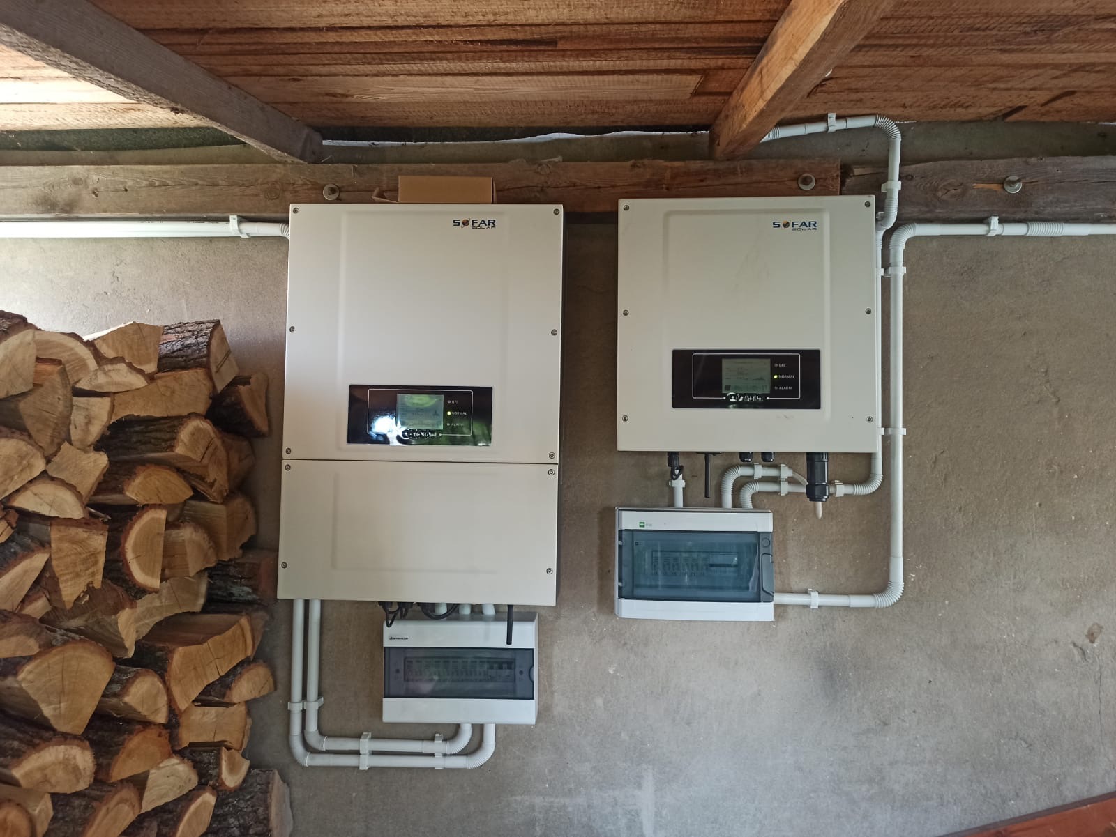 Dwa białe inwertery SOFAR zamontowane na szarej ścianie wewnątrz budynku z drewnianym stropem, obok stos drewna opałowego.