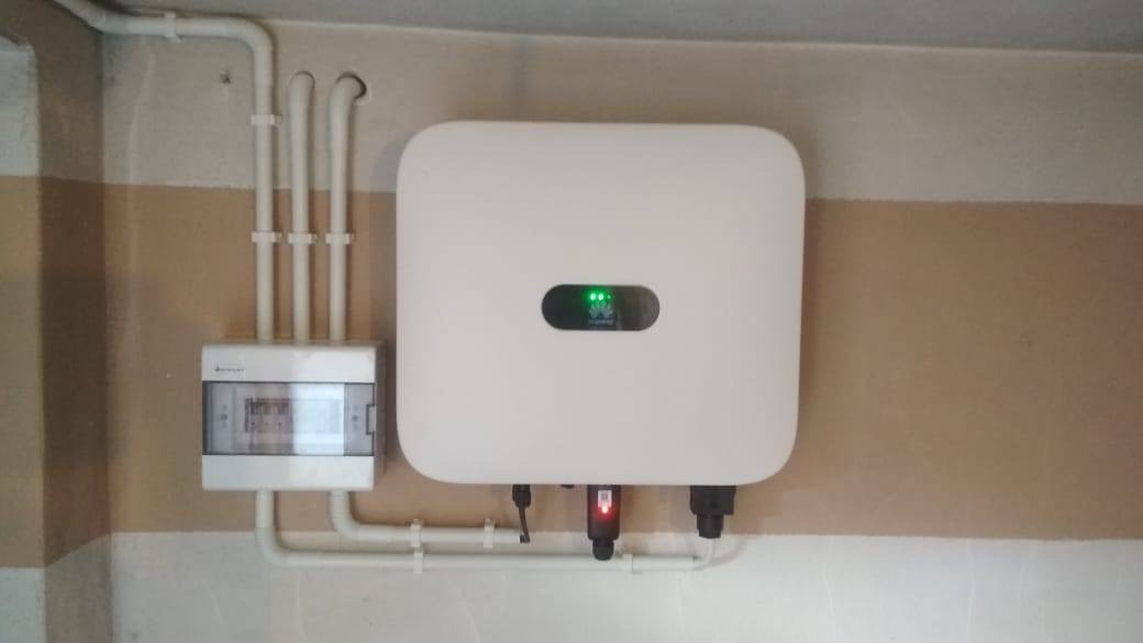 Zamontowany, biały inwerter Huawei z podłączeniami kablowymi i widoczną skrzynką bezpiecznikową na ścianie z pasami w kolorze kremowym i białym.